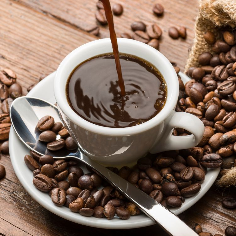 Foto de mesa com grãos de café, colher, pires e café sendo derrubado em xícara branca 