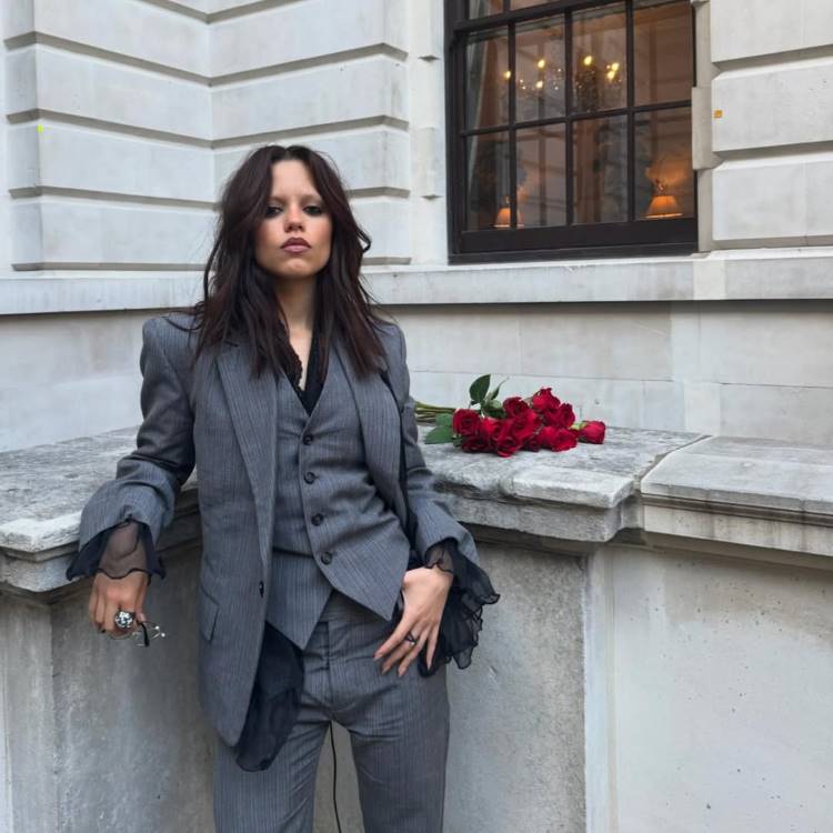 Jenna Ortega veste um terno de três peças cinza. O casaco tem ombros pontudos e mangas de renda preta saem das mangas. Um buquê de rosas vermelhas está sobre um muro de pedra ao lado dela.








