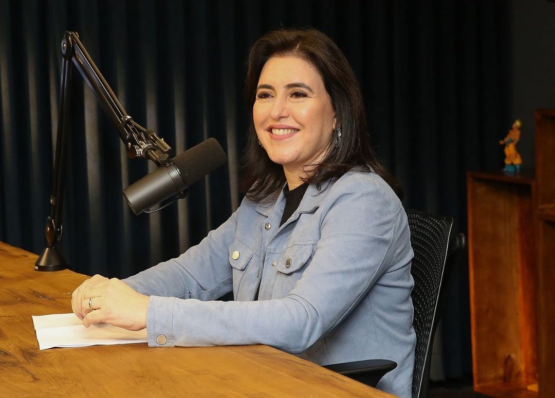 Foto da candidata à presidência nas eleições 2022 Simonte Tebet sentada em uma mesa de entrevista e sorrindo.