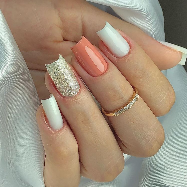A imagem mostra uma mão com unhas decoradas de maneira elegante e variada. Duas unhas são pintadas de branco com pontas arredondadas, uma é de glitter dourado, e outra em um tom de coral brilhante. O design é complementado por um anel dourado com pequenos brilhos. Este conjunto é um exemplo de unha decorada simples, que combina diferentes texturas e cores de forma harmoniosa.