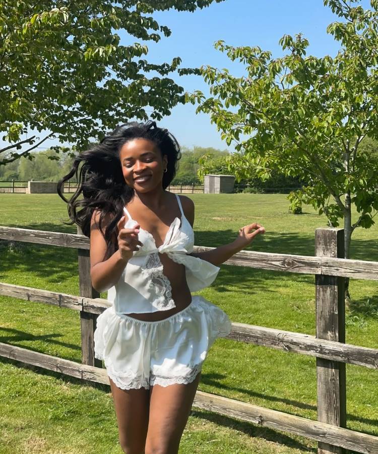Uma mulher sorridente com cabelos longos e escuros, vestindo um conjunto de seda branco com detalhes de renda. Ela corre em um campo verde com uma cerca de madeira e árvores ao fundo, sob o sol.