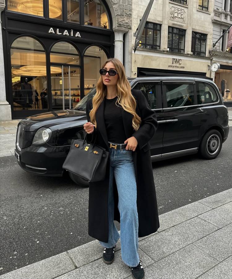 Mulher posa em rua elegante, vestindo sobretudo preto longo, blusa preta básica, jeans de boca larga e tênis escuro. Usa óculos de sol grandes e segura bolsa estruturada preta. Inspiração moderna e prática para looks de trabalho casuais e estilosos.