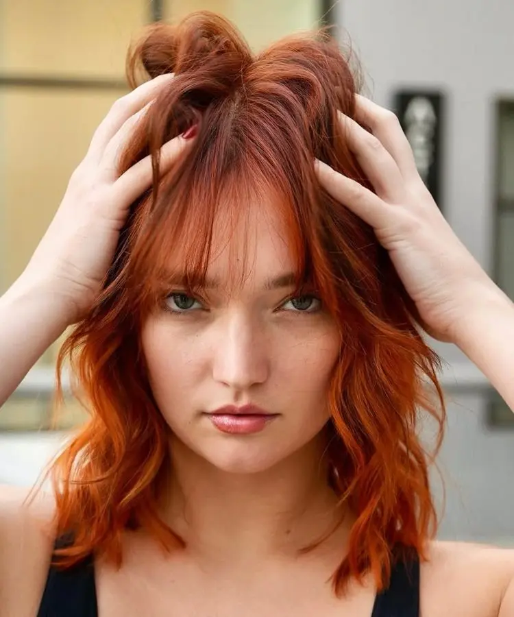 Mulher com cabelo médio ruivo vibrante, corte desfiado e franja leve. Ela segura os cabelos com as mãos, criando um visual descontraído. O fundo desfocado com tons neutros destaca seus traços marcantes e o tom intenso do cabelo.