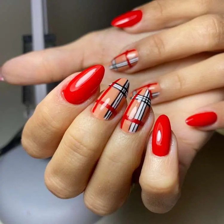 Mão com unhas em formato amendoado. Duas unhas são vermelhas lisas vibrantes. As outras três unhas têm um padrão xadrez em tons de nude, vermelho e preto, simulando o estilo Burberry.