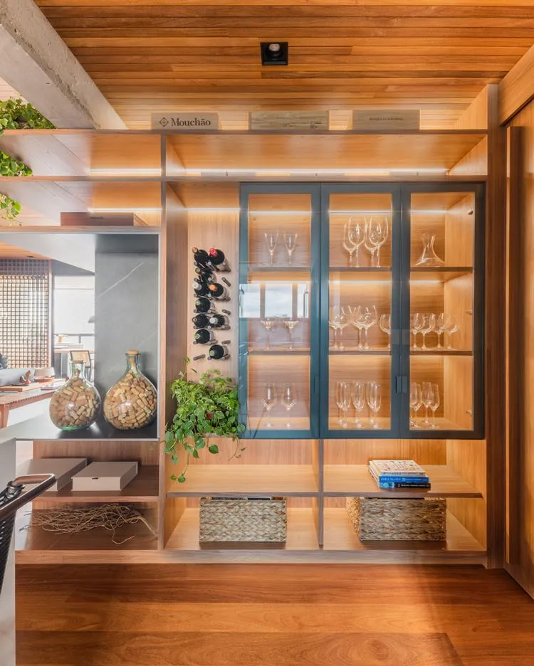 Cristaleira da cozinha de apê de 187 m² decorada pelo arquiteto Pietro Terlizzi, com taças, ao lado pinos que seguram garrafas de bebidas. Caixas de palha, potes de vidro com rolhas, e caixas em MDF nas repartições externas do móvel da cristaleira