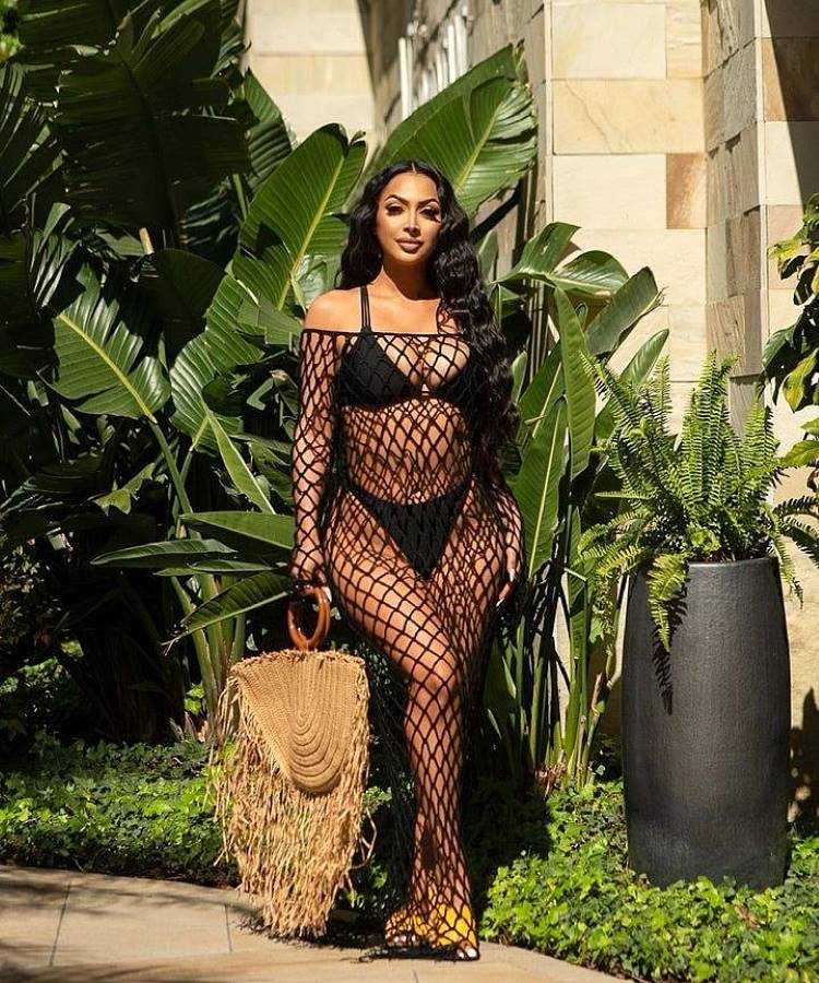 Uma mulher com longos cabelos escuros usa um biquíni preto e uma saída de praia de crochê preta vazada. Ela segura uma bolsa de palha grande com alças de madeira, posando ao lado de plantas tropicais luxuriantes.