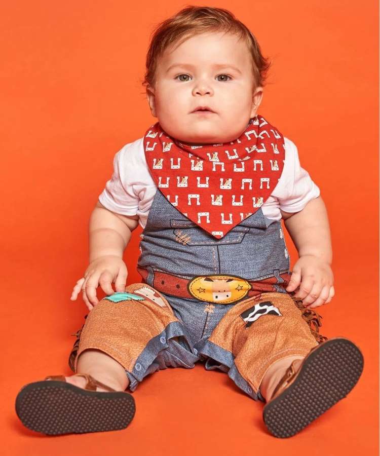 Menino pequeno usando macacão com estampa de menina, camiseta branca, lenço no pescoço e sandalinha.
