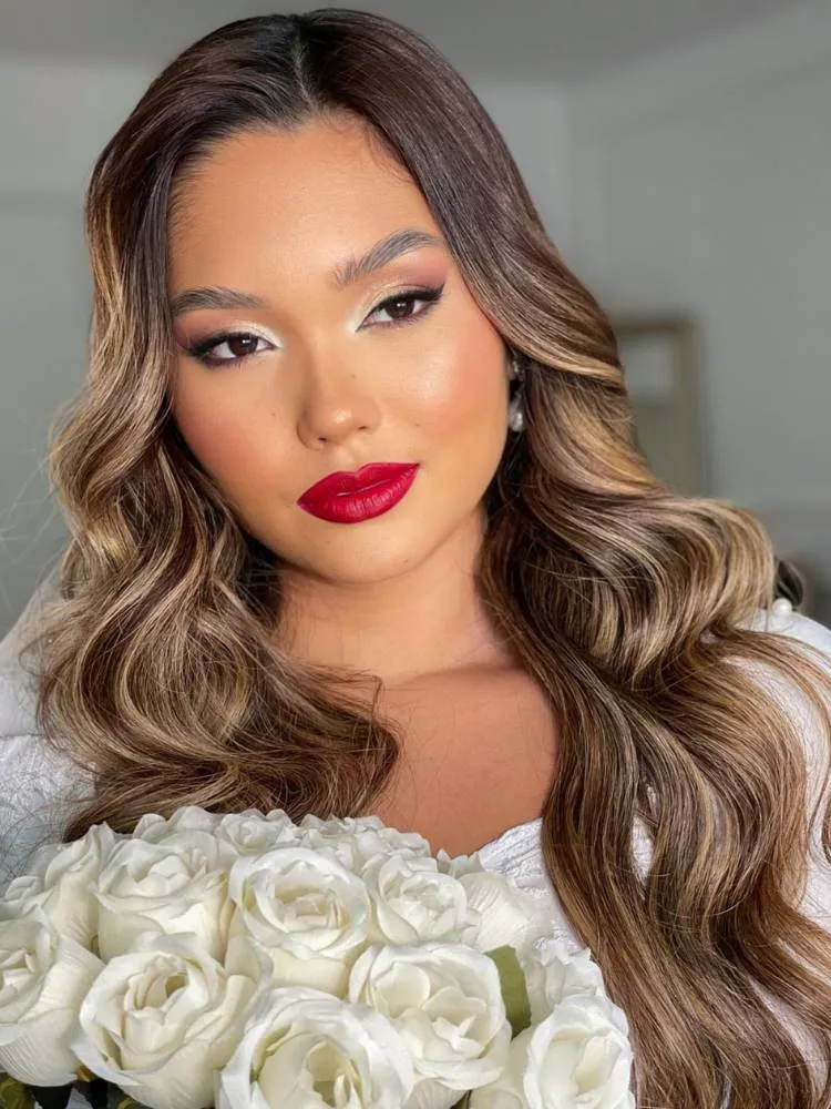 Mulher de pele morena vestida de noiva, segurando buquê de rosas brancas e usando maquiagem natural nos olhos, mas batom vermelho nos lábios 