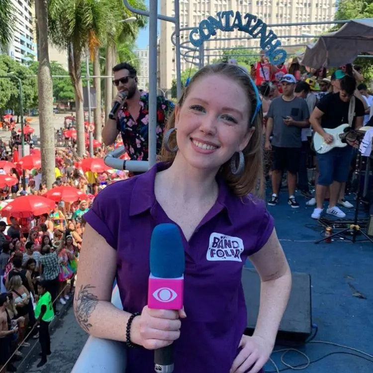 Raquel real em bloquinho de Carnaval, usando polo roxa e tiara escrita "contatinho", segurando microfone da Band