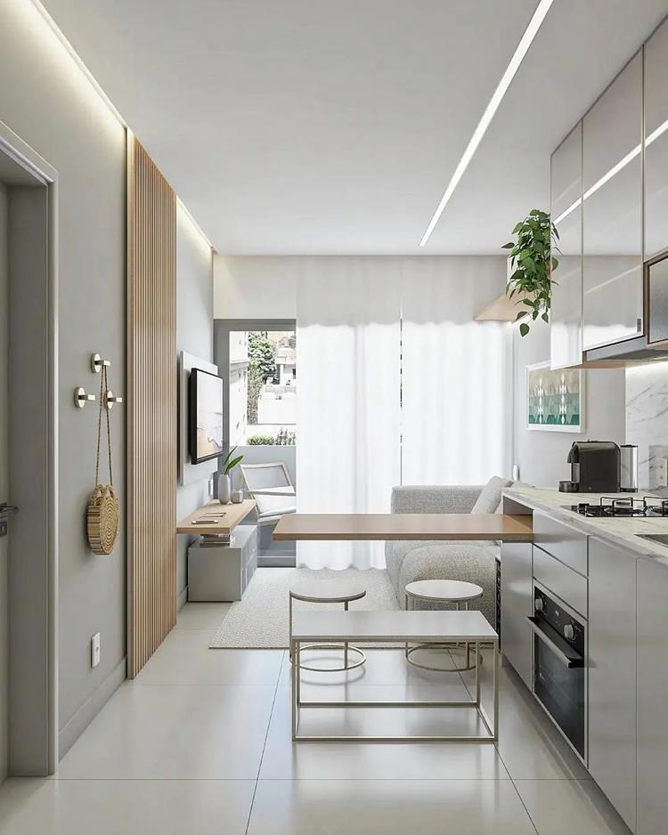 foto de cozinha e sala integrados em apartamento pequeno em tons claros, com brancos, espaço para pendurar bolsas, janela grande, TV em painel, sofá cinza e mais.  O diferencial é uma mesa saindo de um móvel da cozinha.