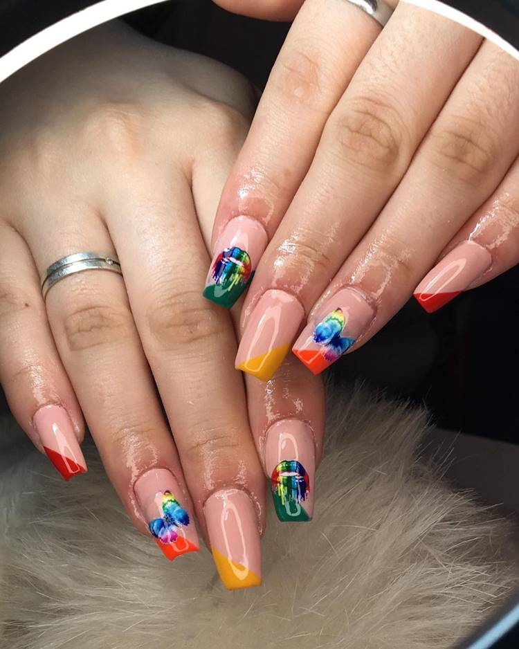 Mão com unhas decoradas coloridas: Polegar não aparece: Indicador, nude com lado diagonal verde e adesivo de boca colorida. Médio, nude com lado diagonal amarelo. Anelas, nude com diagonal laranja e adesivo de borboleta colorida. Mindinho: nude com diagonal vermelho
