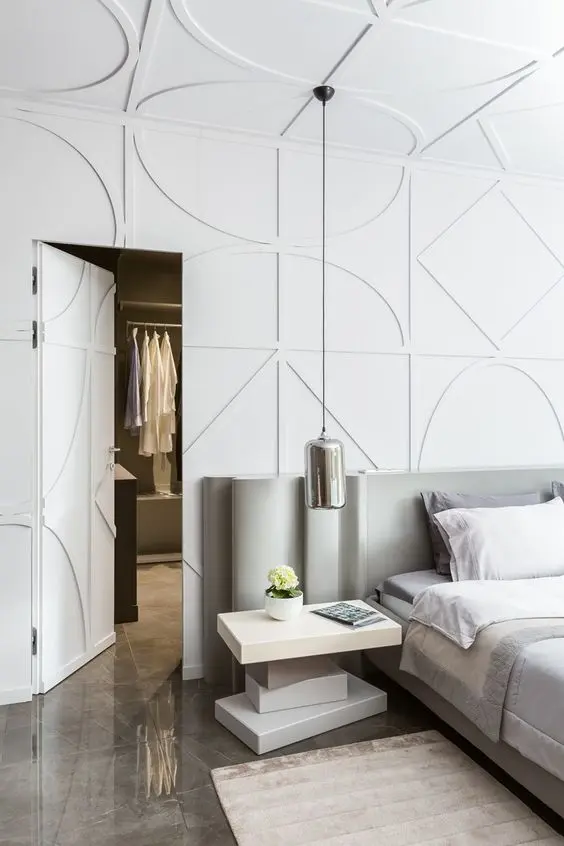 Quarto com parede branca com molduras em formato de formas geométricas, cama, mesa de cabeceira e porta do closet aberta. 