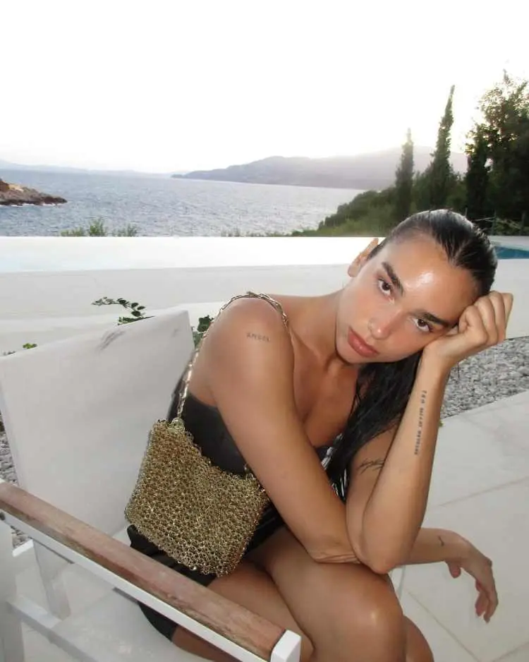 Dua Lipa sentada apoiando a cabeça na mão, veste vestido de alça preto, atrás piscina e mar, exibindo tatuagens nos pulsos, ombro e braço