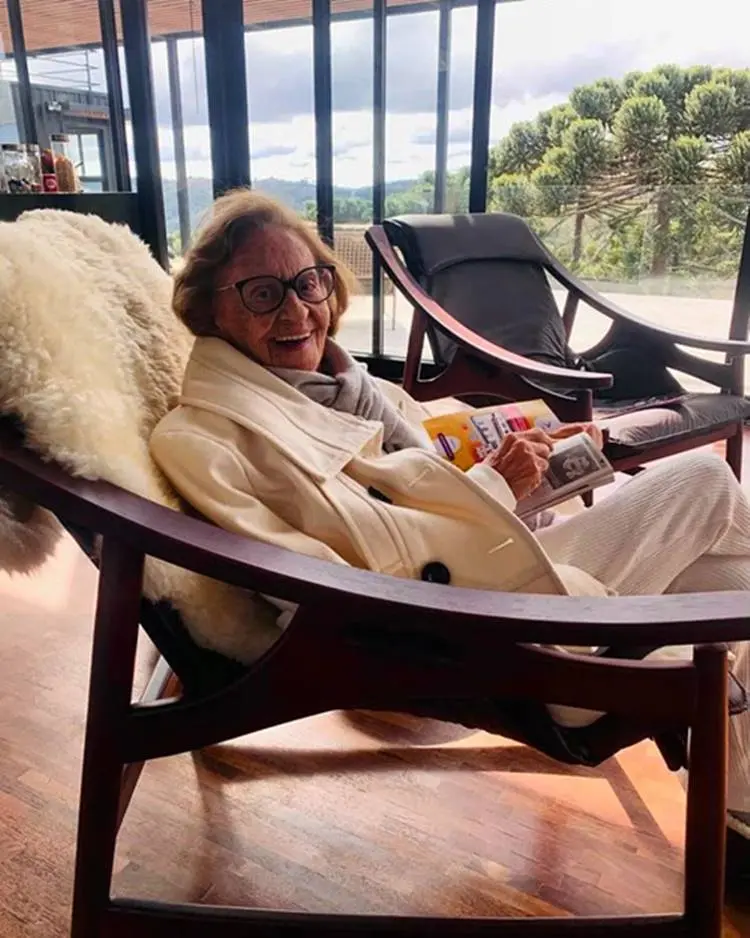 A atriz de óculos com revista na mão sentada em cadeira.