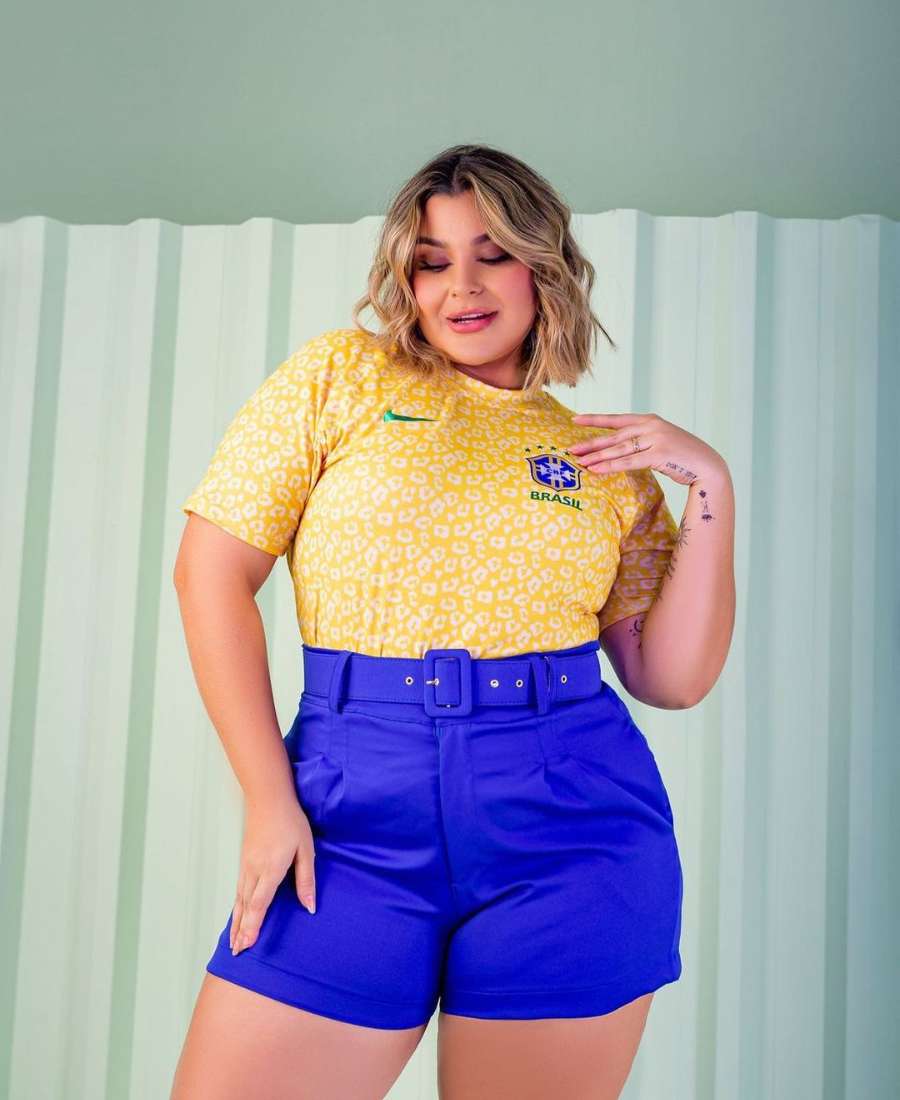 Imagem com fundo de cômodo de casa, com painel branco. No centro, mulher usando roupas para a copa do mundo: camiseta amarela de oncinha do Brasil e short azul.