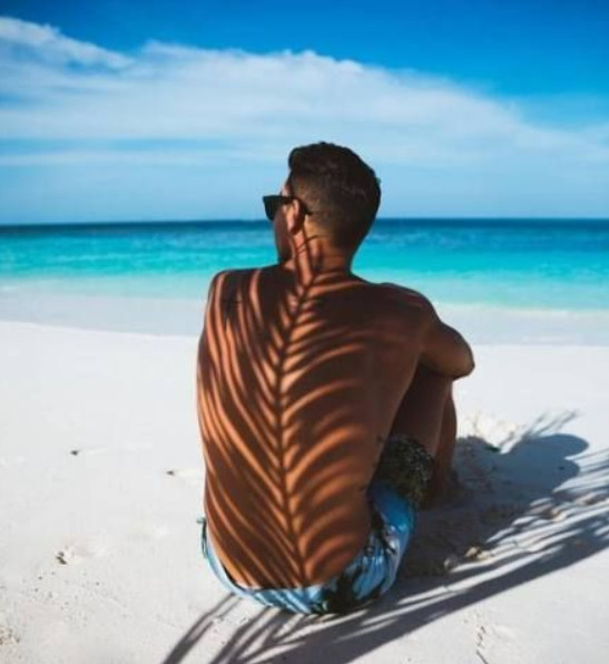Homem de pele clara usando bermuda azul, sentado na areia à beira do mar