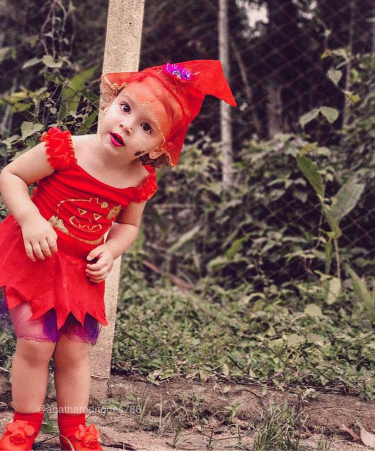 Garota fantasiada de bruxinha.Cores predominantes: vermelho. Usa: vestido, chapéu, sandália e meia