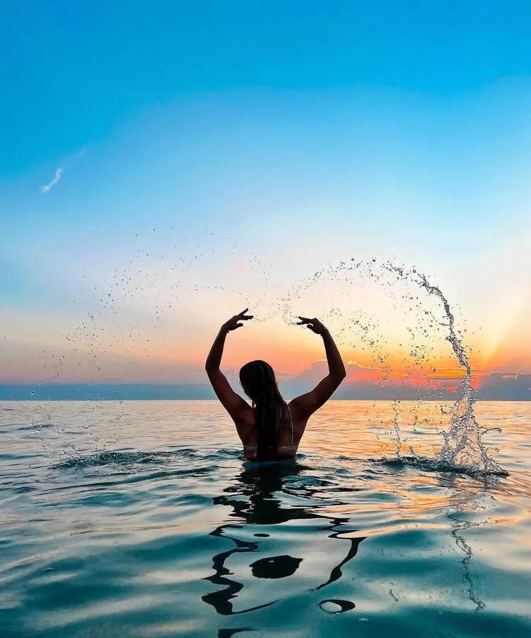 Uma mulher de costas, com cabelos longos e soltos, está no mar ao pôr do sol. Ela levanta os braços, criando um arco de água ao seu redor. O céu está azul e alaranjado. Fotos tumblr na praia frequentemente capturam momentos mágicos como este.


