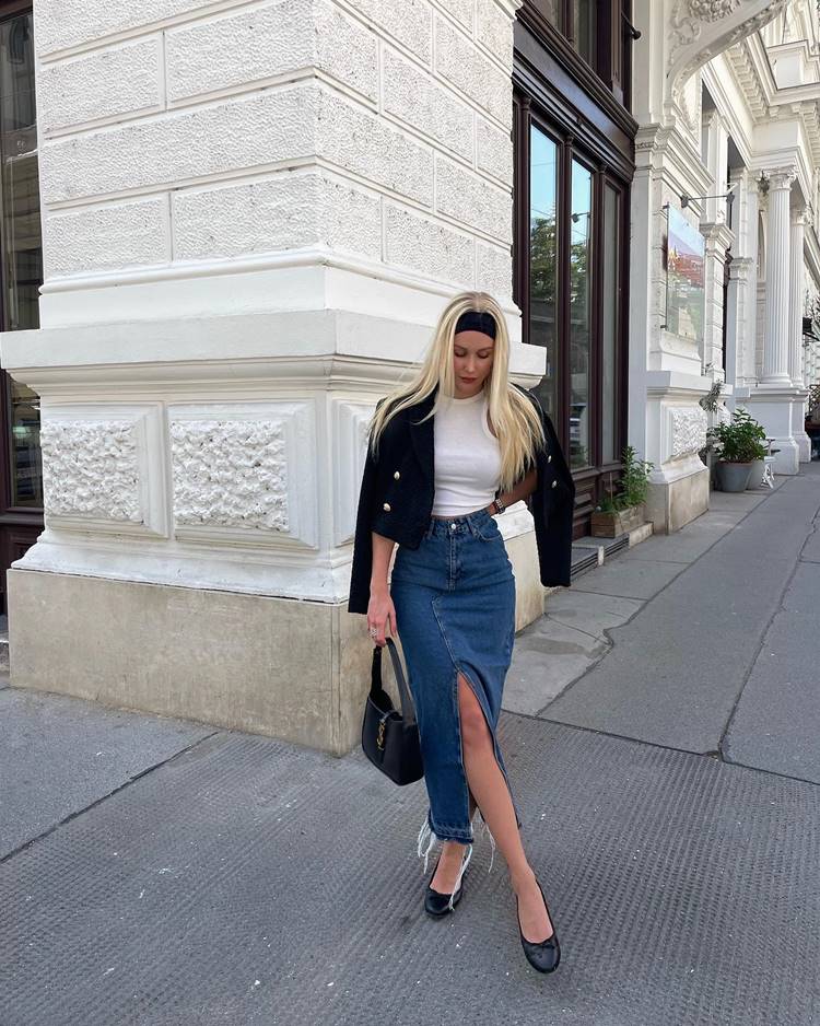 Menina usando regata branca, blazer preto e saia comprida com fenda no joelho, mais sapatilha, bolsa preta e faixa no cabelo, em local público com construção grande.