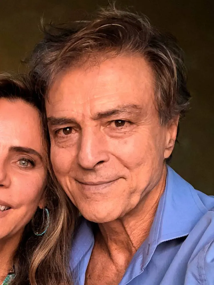 Homem de cabelos grisalhos, levemente despenteados, com expressão serena e sorriso sutil, veste camisa azul clara. Ao lado, parcialmente visível, uma mulher com brincos turquesa. Ex-integrante do elenco de "Vale Tudo", hoje lembrado por seu carisma icônico.