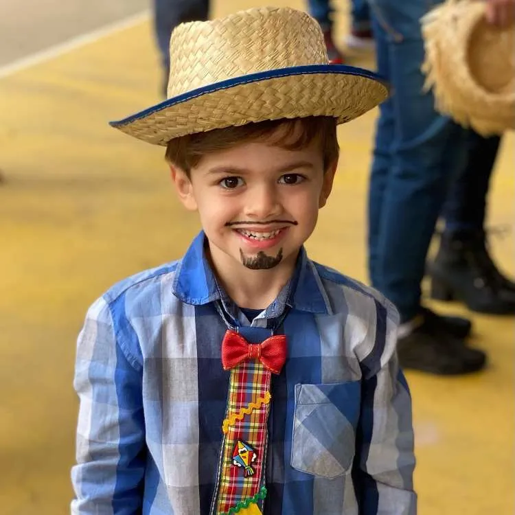Menino usando camisa xadrez azul com gravata decorada e chapéu de palha para festa junina.
