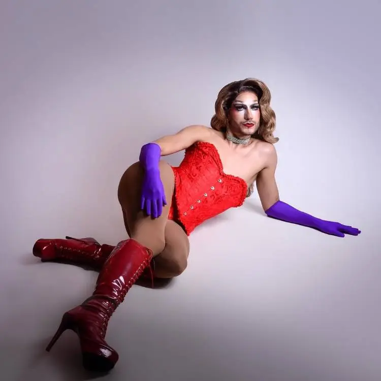 Drag Queen posa em estúdio vestindo corset vermelho e botas de salto combinando. Luvas roxas chamativas e colar prateado completam o visual. Com maquiagem artística intensa, corpo reclinado no chão, a cena transmite ousadia e teatralidade típica de performances drag.