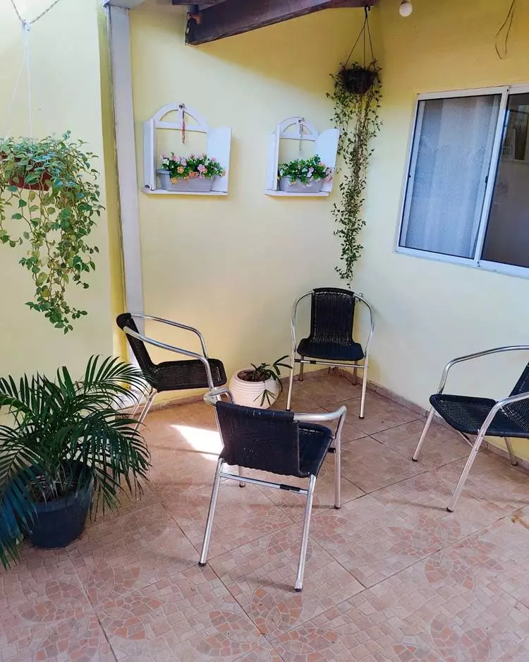 Imagem de fachada de casa simples de cor amarela. Decorada com plantas ´penduradas, em vasos e 4 cadeiras de metal e plástico preto. Na parede à duas decorações em formatos de janelinha, com vasos de flores coloridas