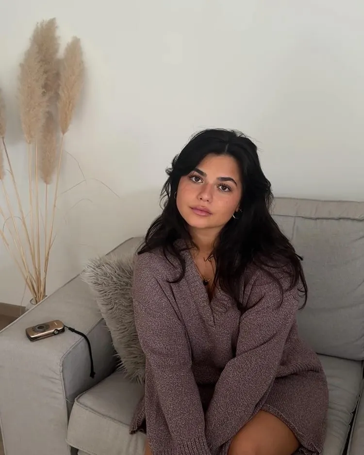 Mulher de rosto redondo com corte cabelo longo usando curtain bangs. Ela usa vestido de lã e está sentada em sofá cinza