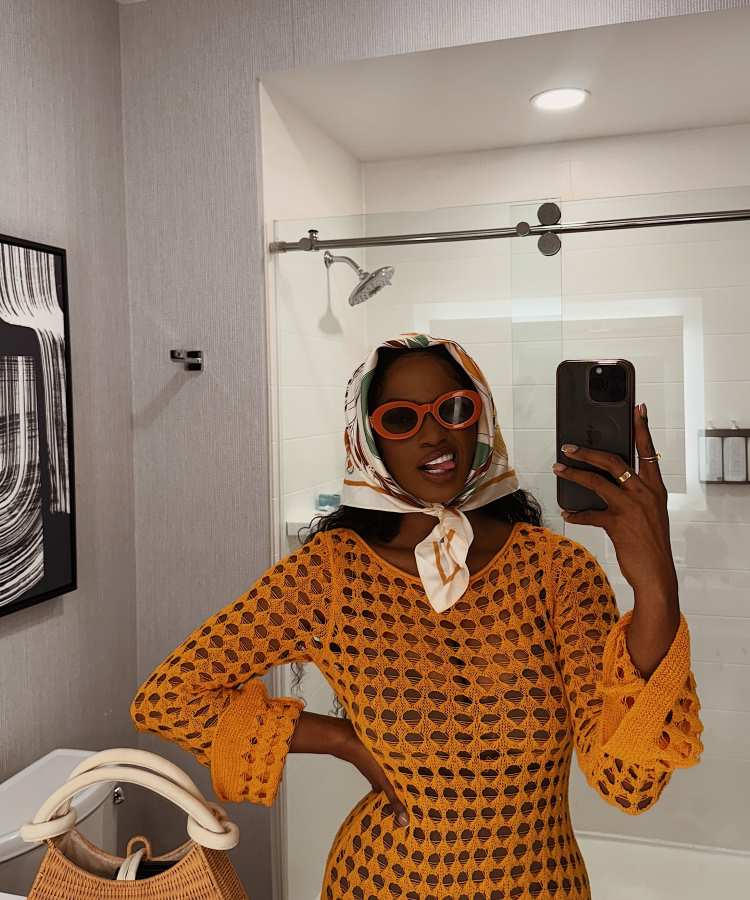 Mulher com cabelo coberto por lenço estampado, óculos laranja, vestido de crochê amarelo, tirando selfie em banheiro moderno.