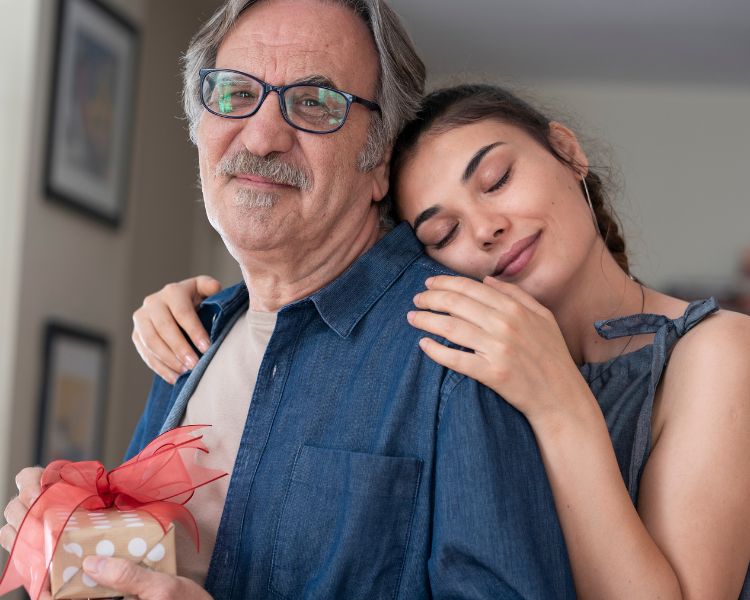 Filha de pele clara abraçando pai, também de pele clara, que segura um presente nas mãos