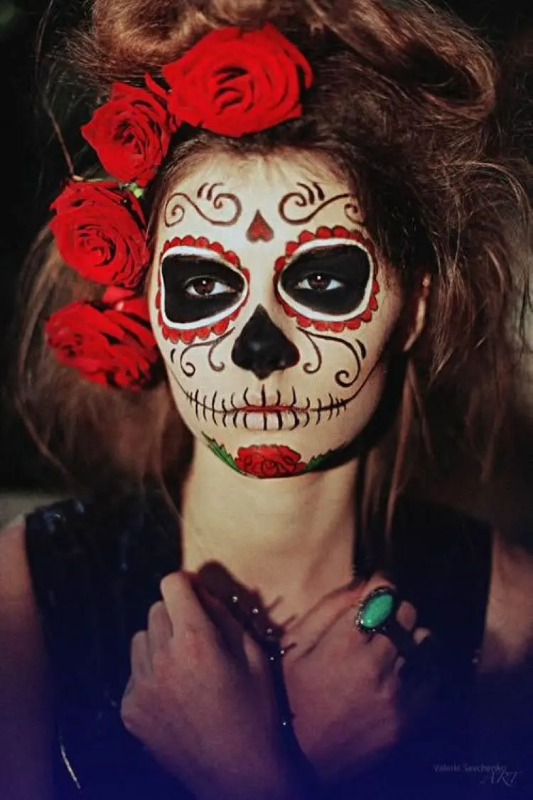 Mulher de pele clara usando maquiagem de caveira mexicana em preto e vermelho e flores vermelhas no cabelo