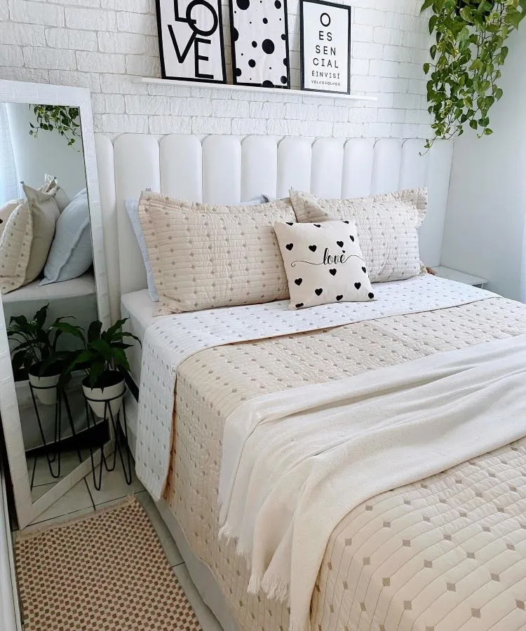A imagem mostra uma decoração fácil para quarto com uma cama de cabeceira branca e roupa de cama em tons neutros. Há um quadro com texto na parede, plantas verdes que adicionam um toque natural e um tapete texturizado. O ambiente transmite uma sensação de tranquilidade e conforto, ideal para pessoas que buscam simplicidade e elegância no design do quarto.

