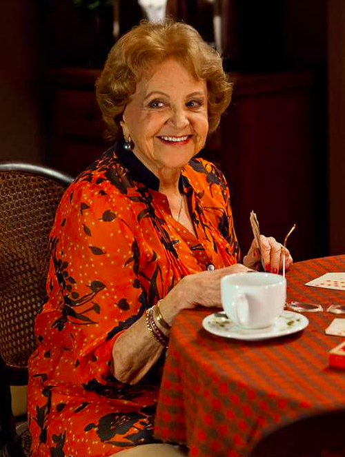 Eva Todor de camisa vermelha e preta sentada à mesa jogando baralho