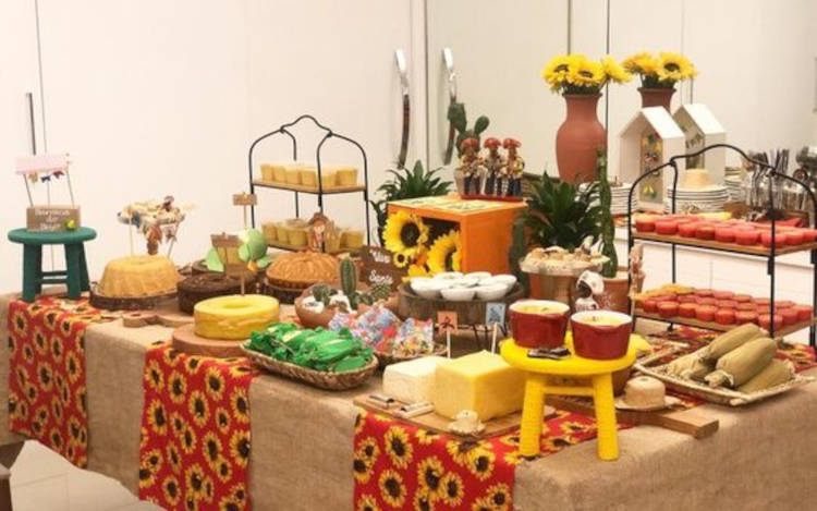 A imagem representa decoração para festa junina. Contém doces e comidas típicas, chapéus de palha, quadros, retalho de girassol