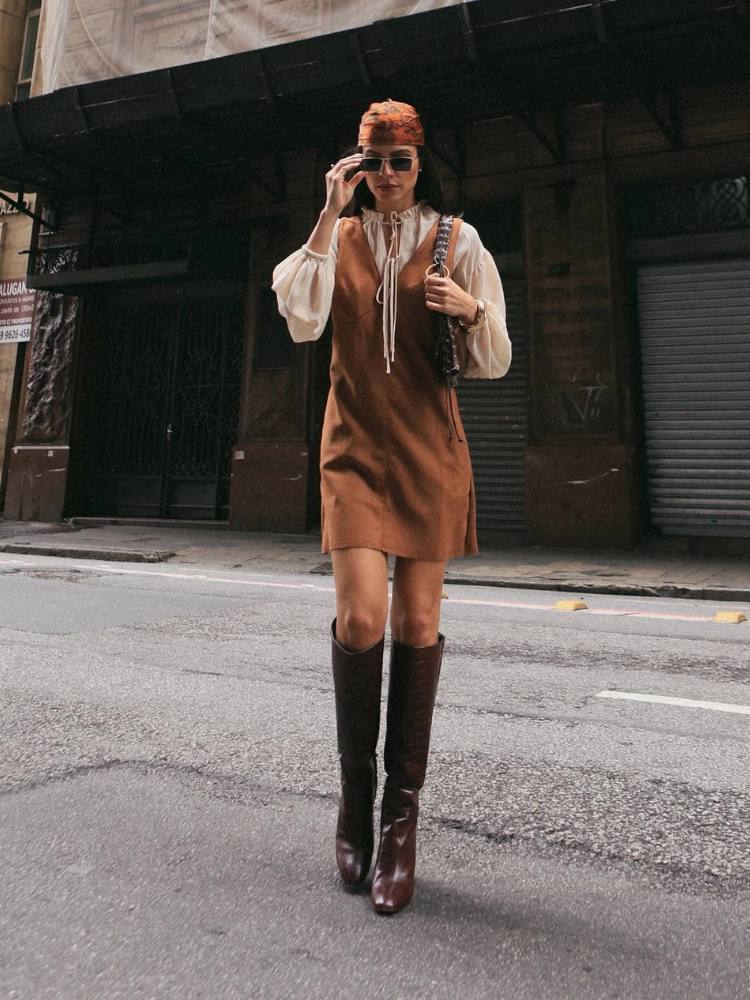 Mulher de pele clara usando camisa bege por baixo de vestido marrom de camurça de bota cano alto 