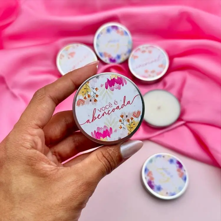 Lata decorativa com tampa estampada de flores coloridas e a frase "Você é abençoada", contendo vela artesanal. Fundo com tecido rosa e outras latas ao redor, ideal como lembrancinha delicada e temática para comemorações especiais, como o Dia da Mulher.