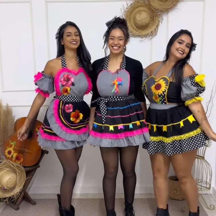 Três mulheres sorridentes vestem aventais juninos coloridos. Dois aventais são rosa choque e xadrez, um com flores e outro com fitas. O terceiro é preto com girassóis e babados amarelos. Todos têm detalhes de bolinhas e saias rodadas.


Fontes





