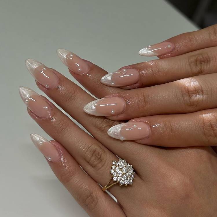 Mãos femininas com unhas longas em formato amendoado, decoradas com esmalte rosa nude, pontas francesas brancas e detalhes em pérolas formando corações. Tatuagens sutis adornam os dedos. Uma malha bege ao fundo cria um cenário aconchegante e elegante.