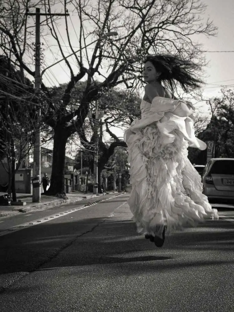 Mulher não-modelo com mais de 30 anos em ensaio fotográfico, usando vestido de babados, correndo por rua. Foto preto e branco