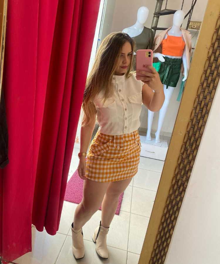 Selfie no espelho mulher com blusa branca, saia xadrez e bota clara