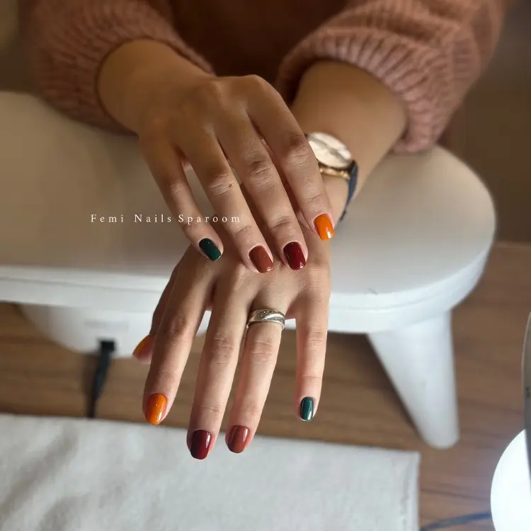 Mãos de pessoa de peel clara em unhas curtas com tons como laranja, marrom, vinho e verde 