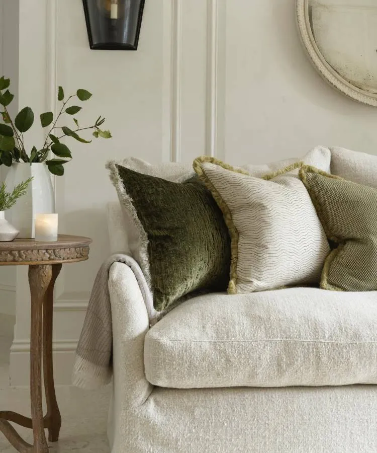 Uma sala elegante com sofá claro e almofadas para sala em tons de verde e creme, com texturas e franjas. Ao lado, uma mesa de madeira redonda exibe um vaso com folhagens e uma vela acesa. Um espelho redondo na parede e uma luminária preta completam o ambiente sofisticado.







