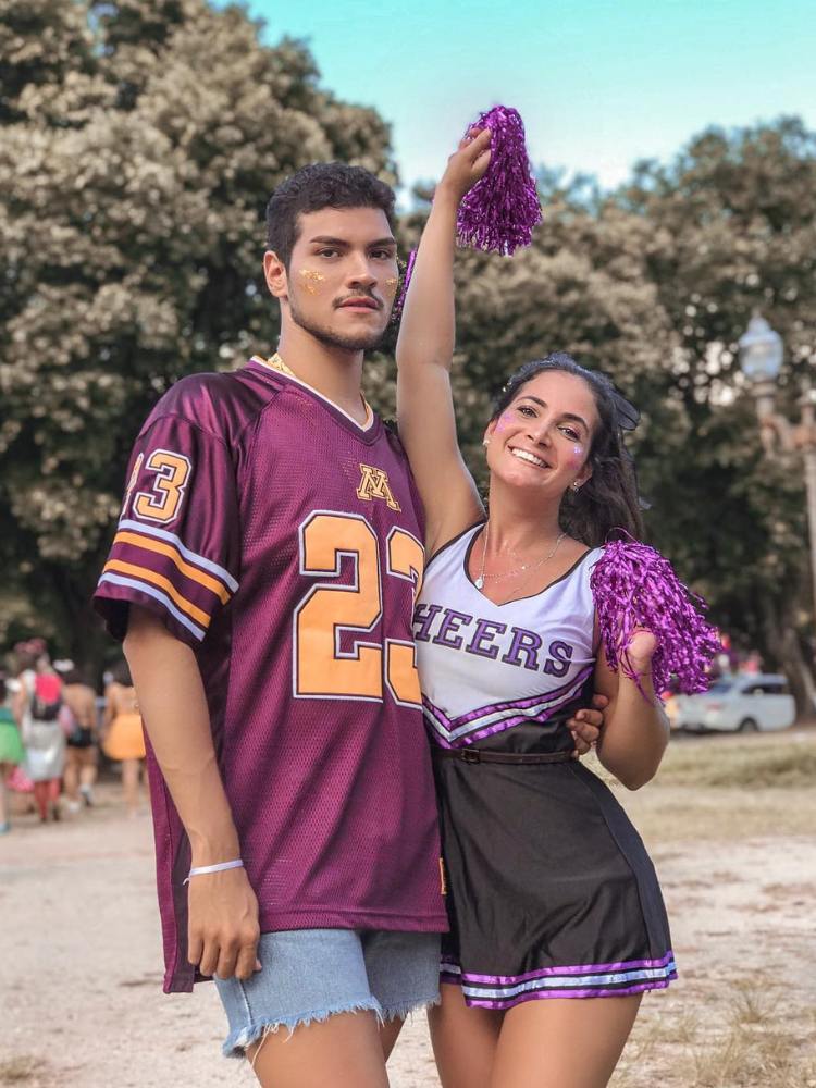 Homem de pele clara com camisa roxa de time e mulher de pele clara com uniforme de cheerleader em roxo, branco e preto