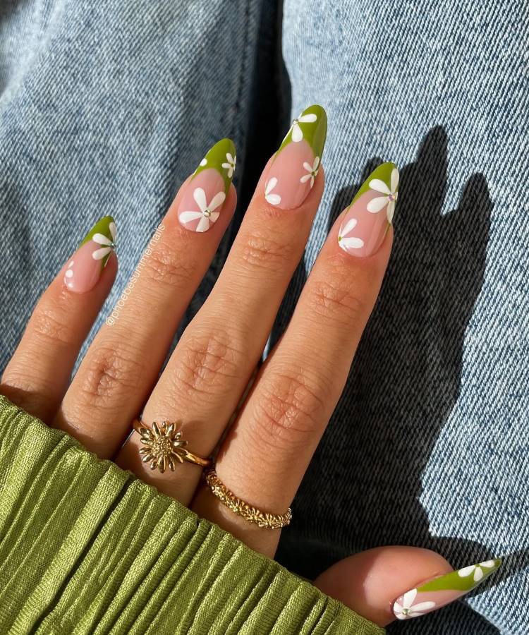 A imagem mostra unhas decoradas com pontas verdes e desenhos de flores brancas delicadas, destacando um visual alegre e primaveril. A mão está adornada com anéis dourados e apoiada sobre jeans, complementada por uma blusa verde plissada elegante.