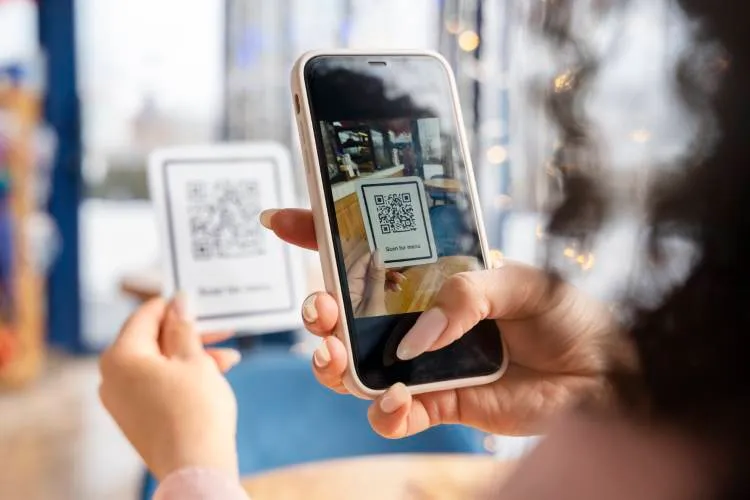 Mulher de unhas feitas segurando uma plaquinha de QR Code com uma mão e um celular lendo o QR Code na outra mão.