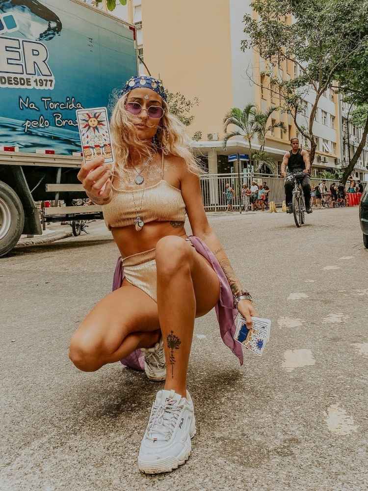 Mulher de pele clara com top e hot pant dourados, acessórios que imitam cigana, lenço na cabeça e cartas de tarot