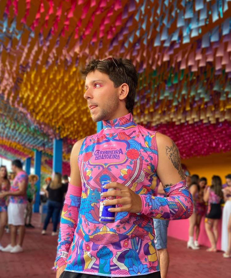 A imagem mostra um homem em um evento carnavalesco, usando um abadá customizado com estampas vibrantes em tons de rosa, azul e amarelo. O abadá possui mangas removíveis, combinando com a decoração colorida ao fundo, cheia de bandeirinhas.