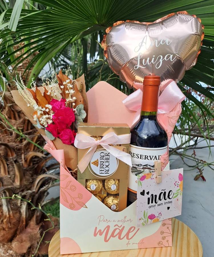 Caixa presente para Dia das Mães com vinho, chocolates Ferrero Rocher, flores e balão dourado em formato de coração. O cartão diz "mãe, amor sem limites". Decoração externa com padrão floral em rosa e laranja. Fundo com folhas verdes.