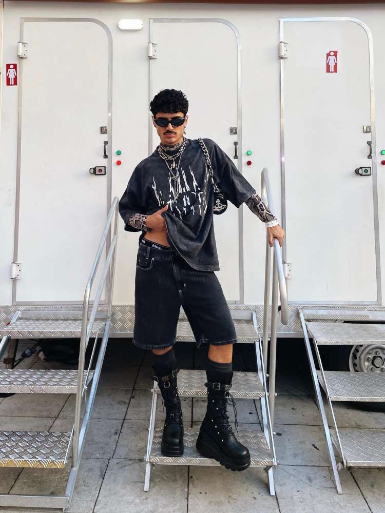 Homem de pele morena usando camisa preta com desenho branco, correntes, bolsa, bermuda preta, óculos de sol e bota