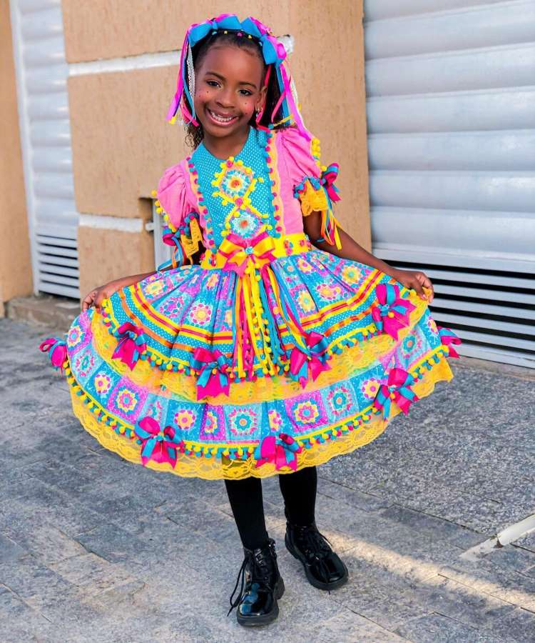 Menina usando vestido de quadrilha colorido, com laços no cabelo, meia-calça preta e sapato preto.
